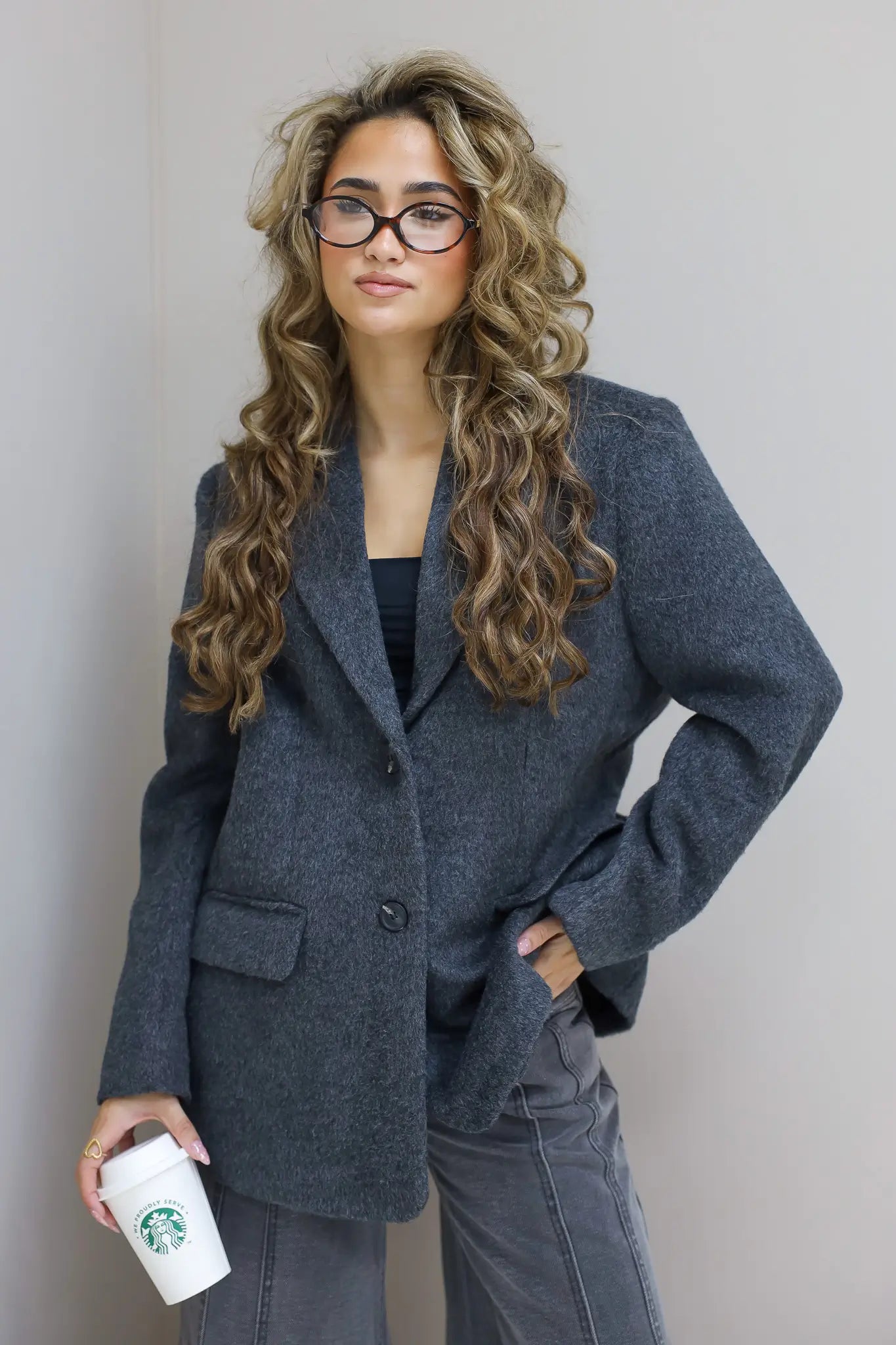 Model draagt de donkergrijze wollen blazer met lange krullen en bril, koffie in hand