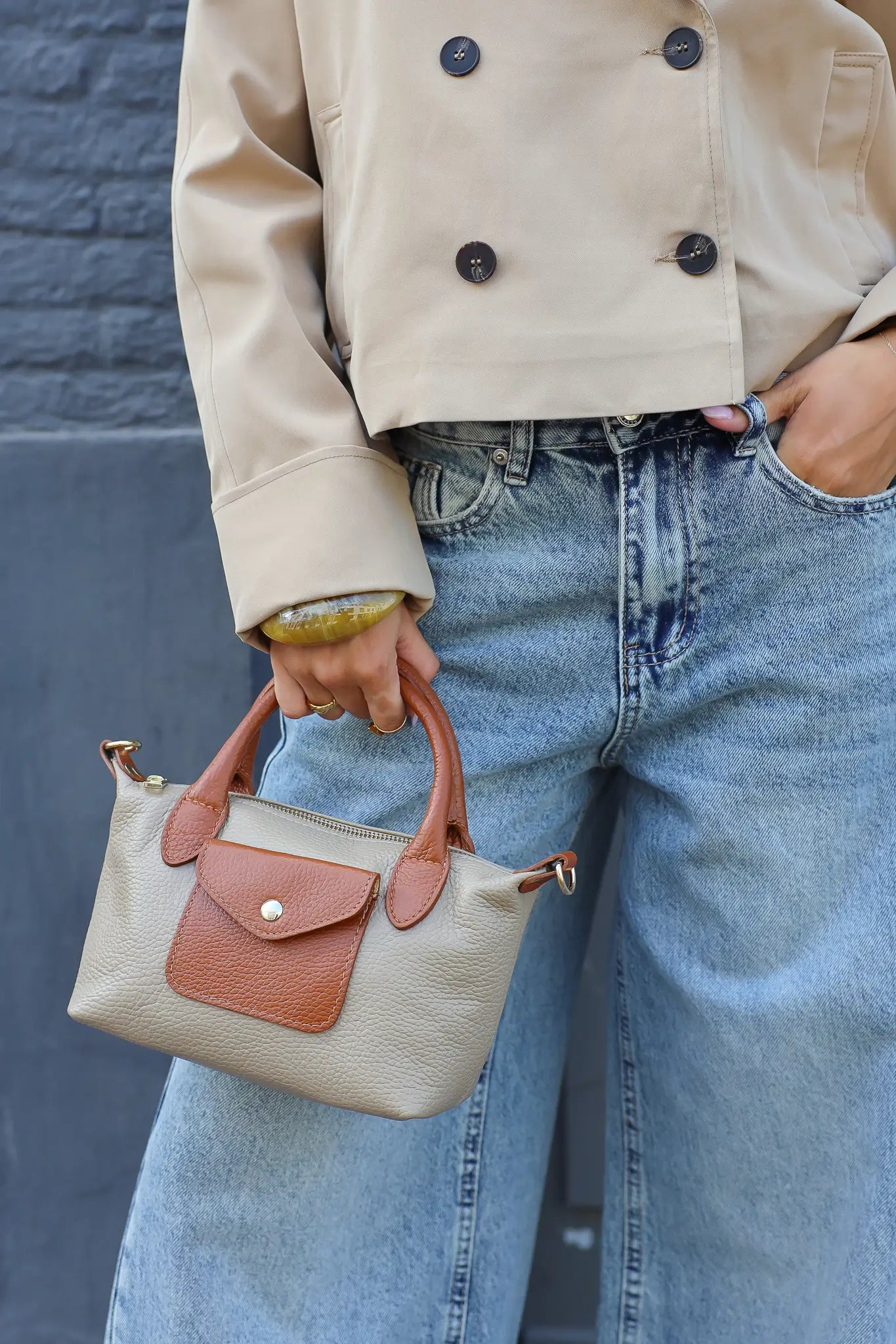beige handtas met bruine leren details vastgehouden bij een jeans en beige jas