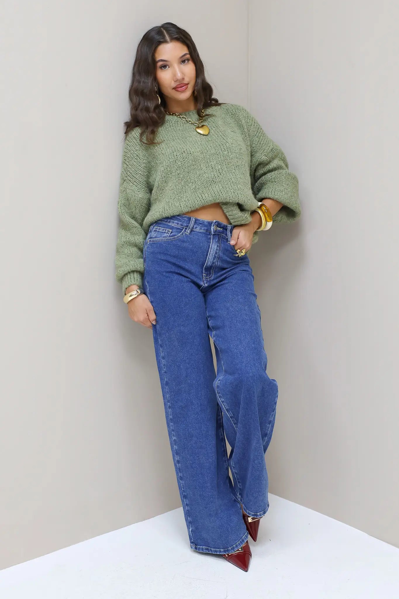 Model draagt blauwe wijde jeans met groene trui en hakken, poserend in hoek van de muur