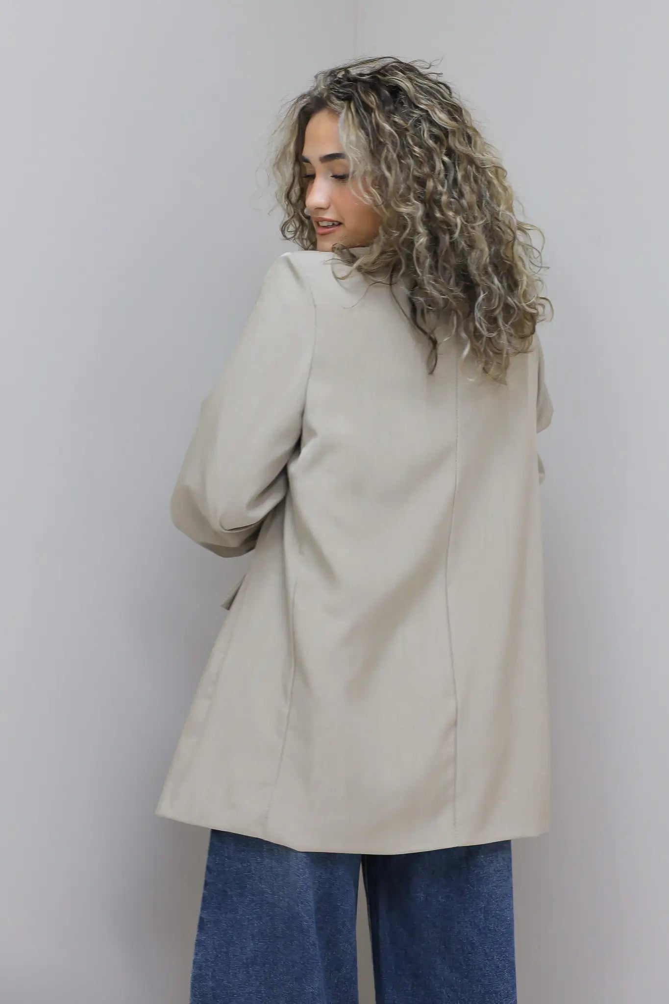 vrouw met beige oversized blazer lacht en poseert ontspannen tegen grijze muur