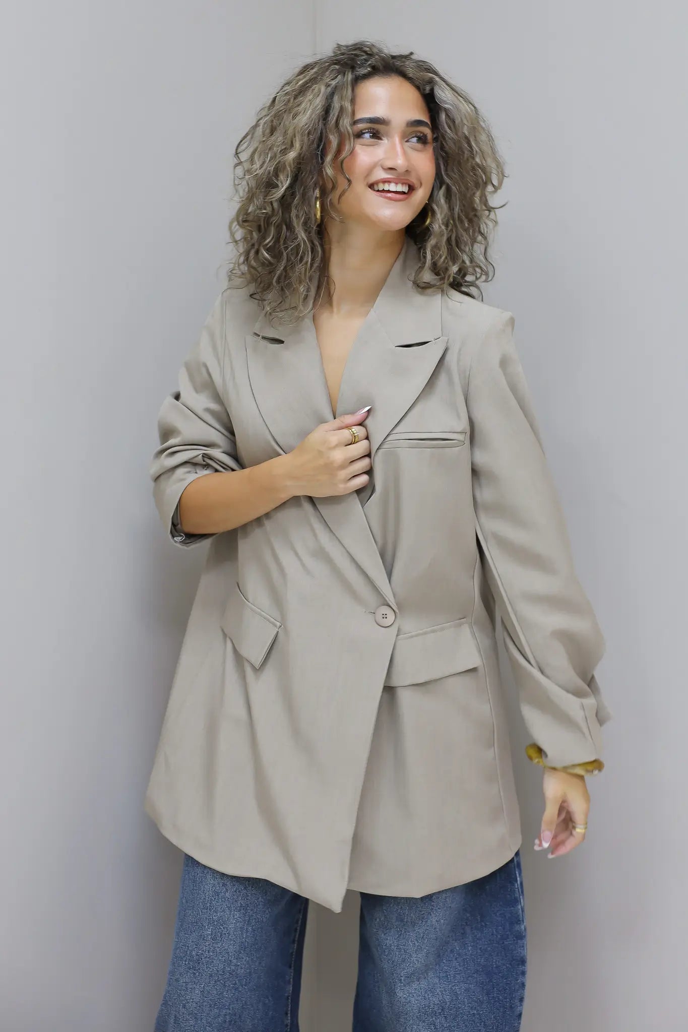 vrouw met krullen draagt beige oversized blazer met jeans en lacht tegen grijze muur