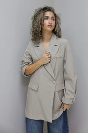 vrouw met beige oversized blazer en jeans kijkt over schouder tegen lichte muur