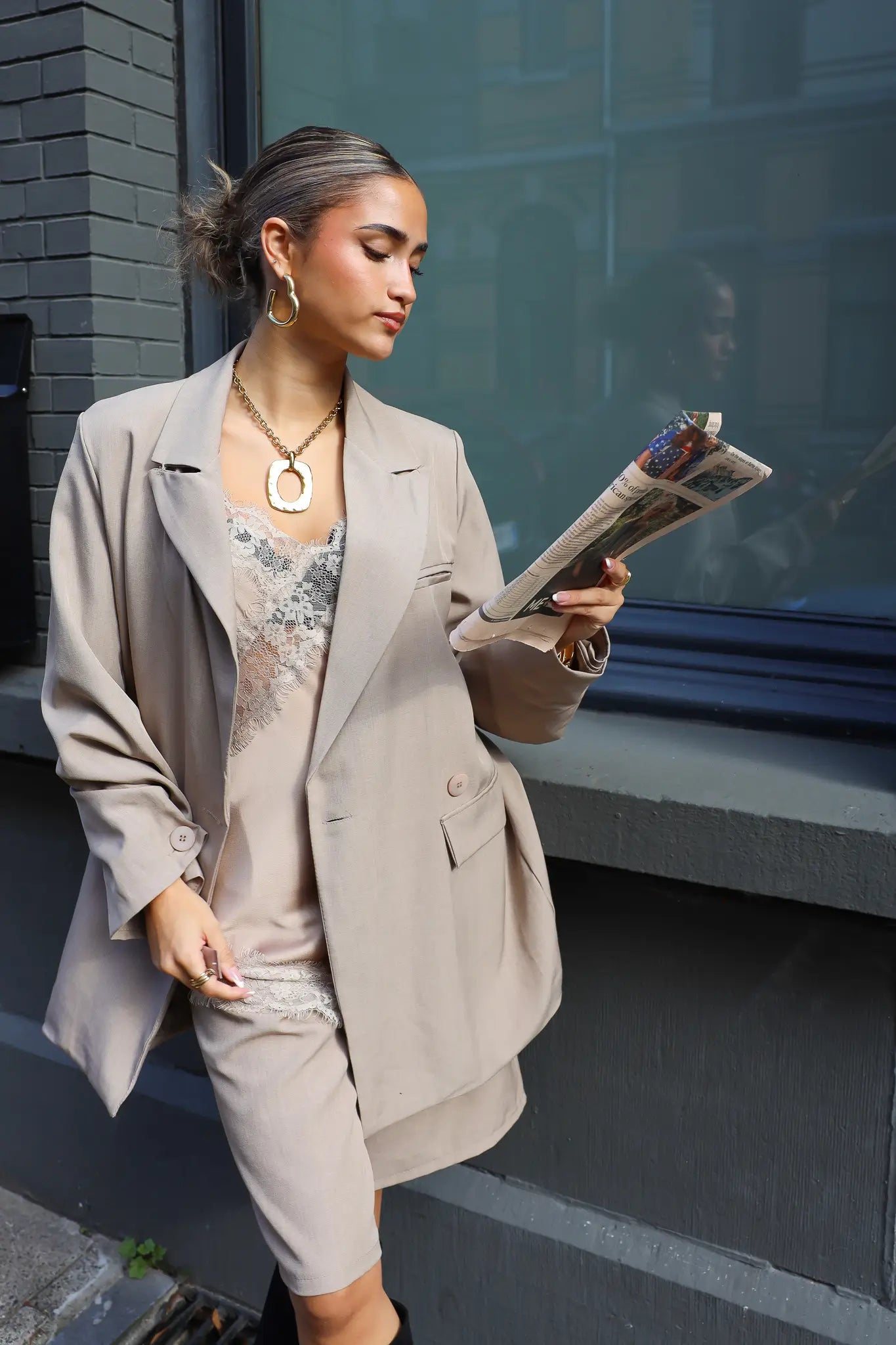 vrouw met beige oversized blazer en zwarte laarzen kijkt op telefoon bij gevel