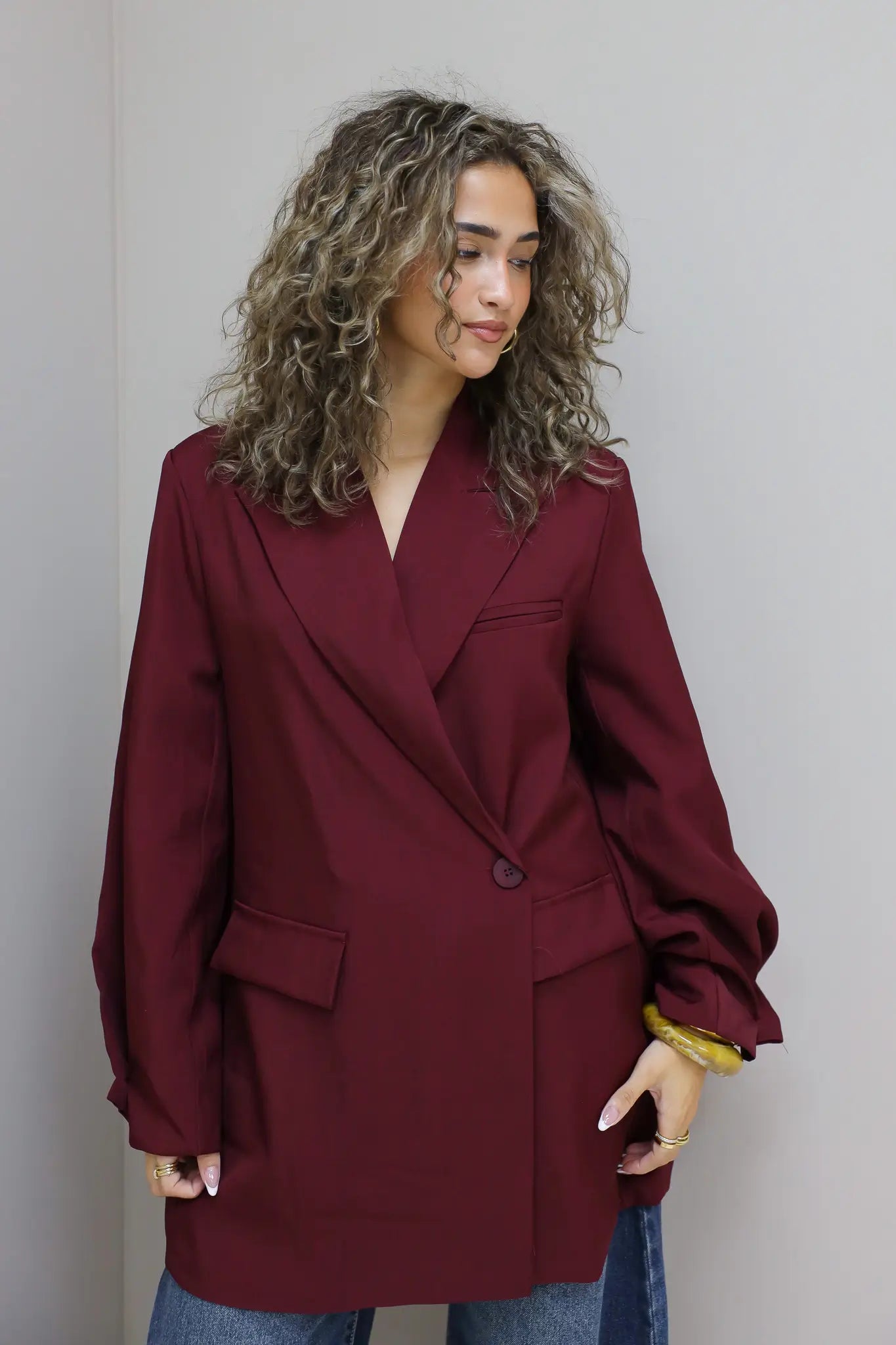 achteraanzicht van vrouw met krullen in bordeaux oversized blazer tegen grijze muur