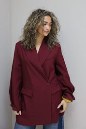 achteraanzicht van vrouw met krullen in bordeaux oversized blazer tegen grijze muur