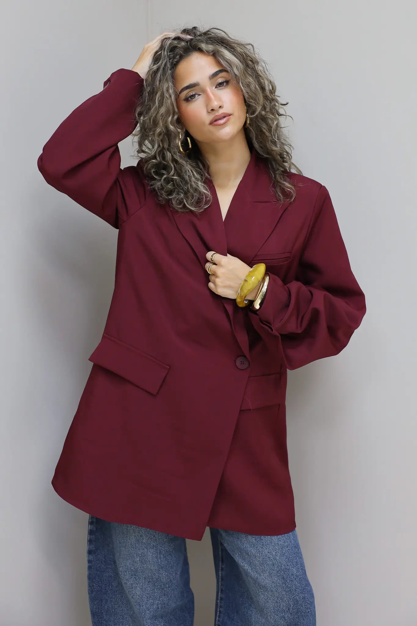 vrouw met krullen draagt bordeaux oversized blazer en jeans tegen grijze muur