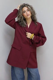 vrouw met krullen draagt bordeaux oversized blazer en jeans tegen grijze muur