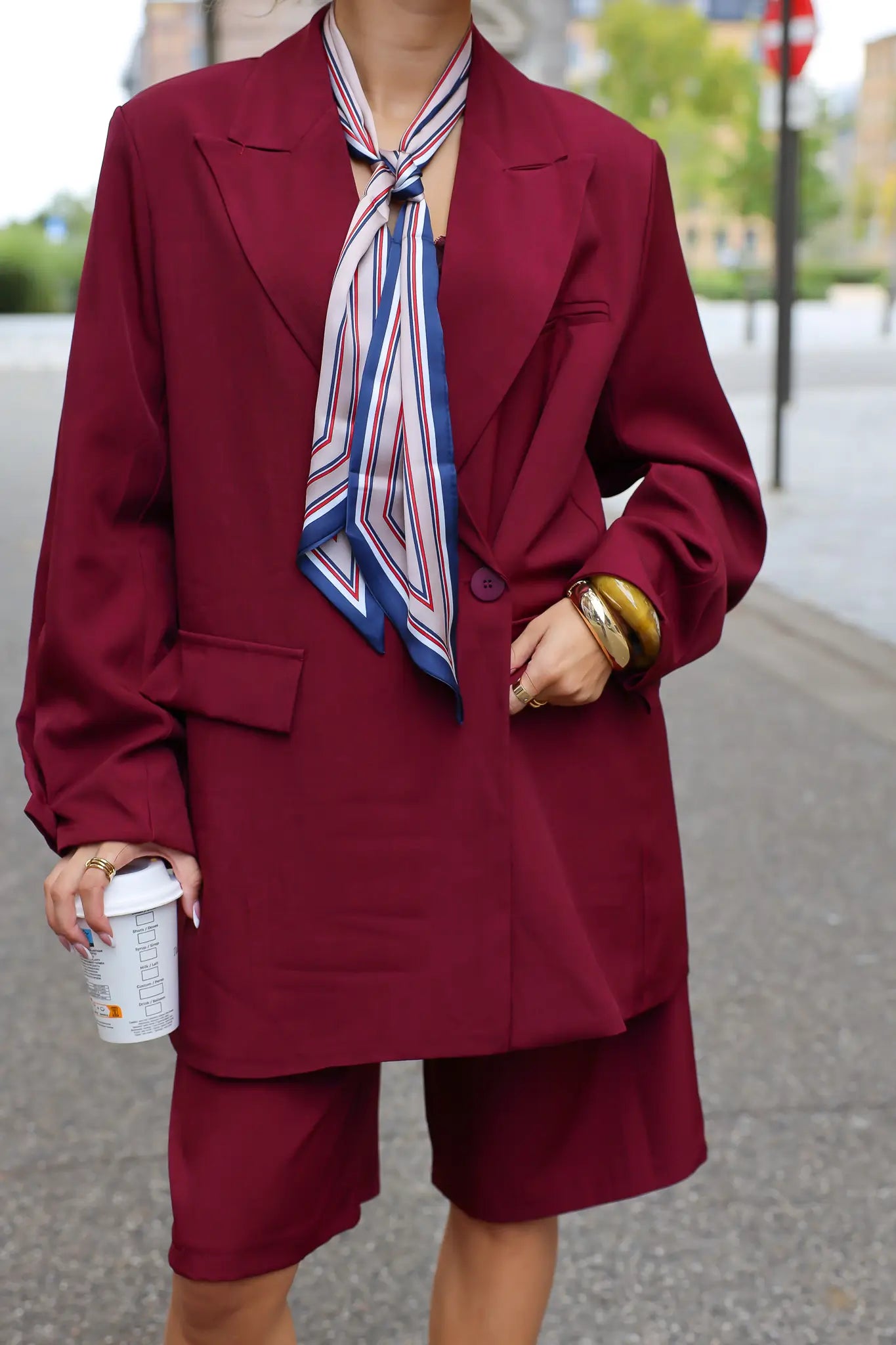 close up van vrouw met bordeaux oversized blazer en geknoopte sjaal bij ingang van gebouw