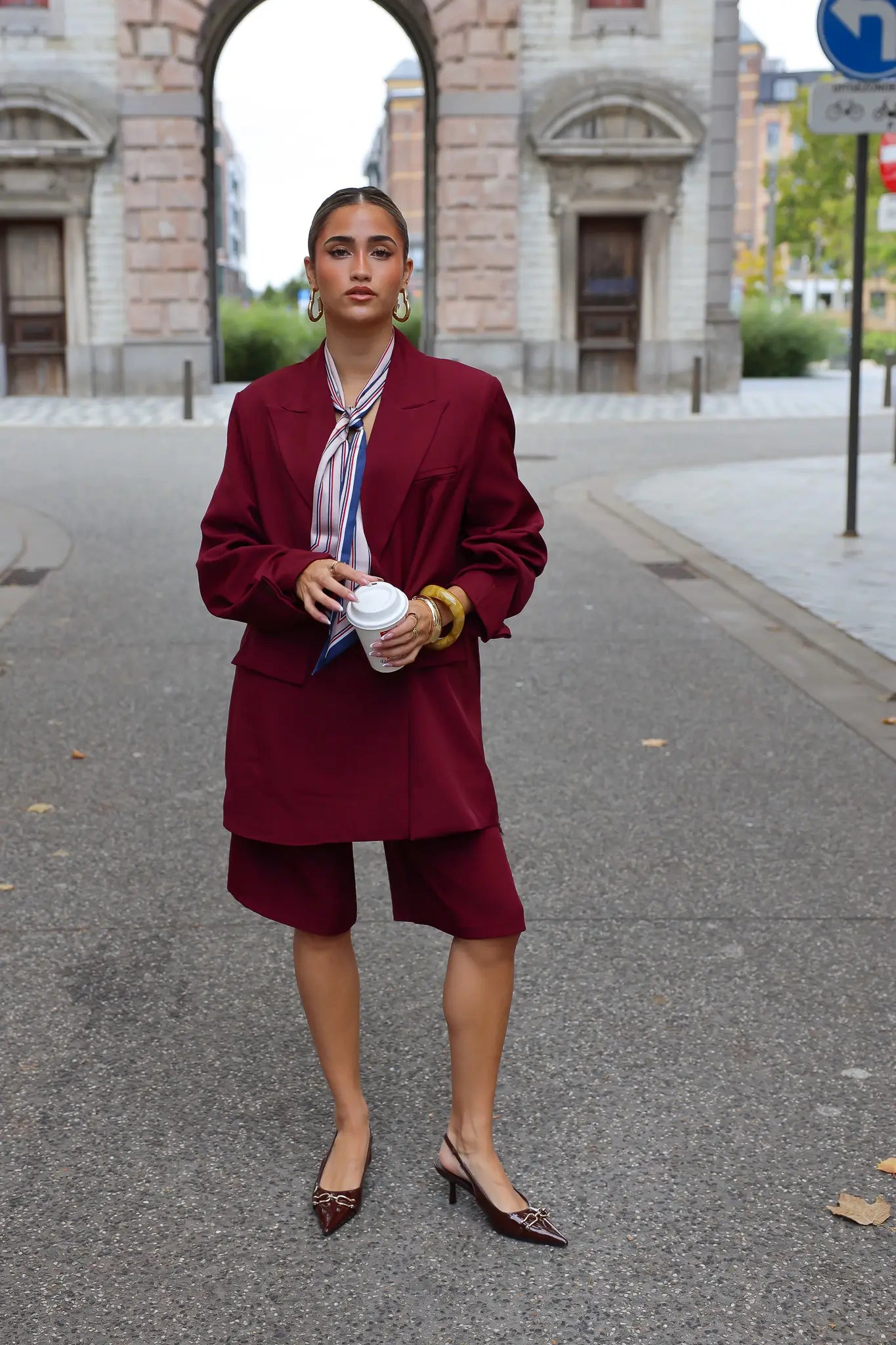 vrouw draagt bordeaux oversized blazer met bijpassende short en hakken op straat bij stadspoort