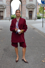 vrouw draagt bordeaux oversized blazer met bijpassende short en hakken op straat bij stadspoort