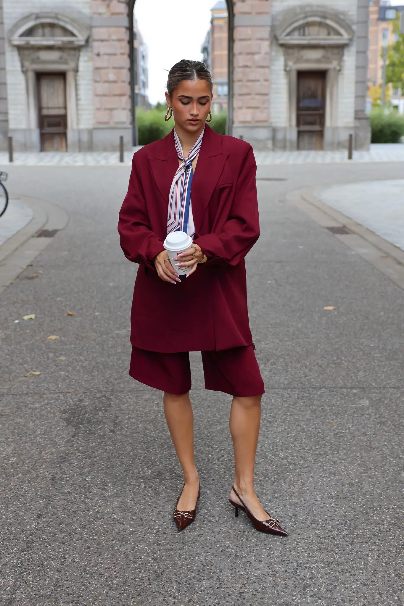 vrouw draagt bordeaux oversized blazer met bijpassende short en hakken recht vooruit kijkend
