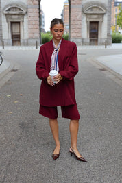 vrouw draagt bordeaux oversized blazer met bijpassende short en hakken recht vooruit kijkend