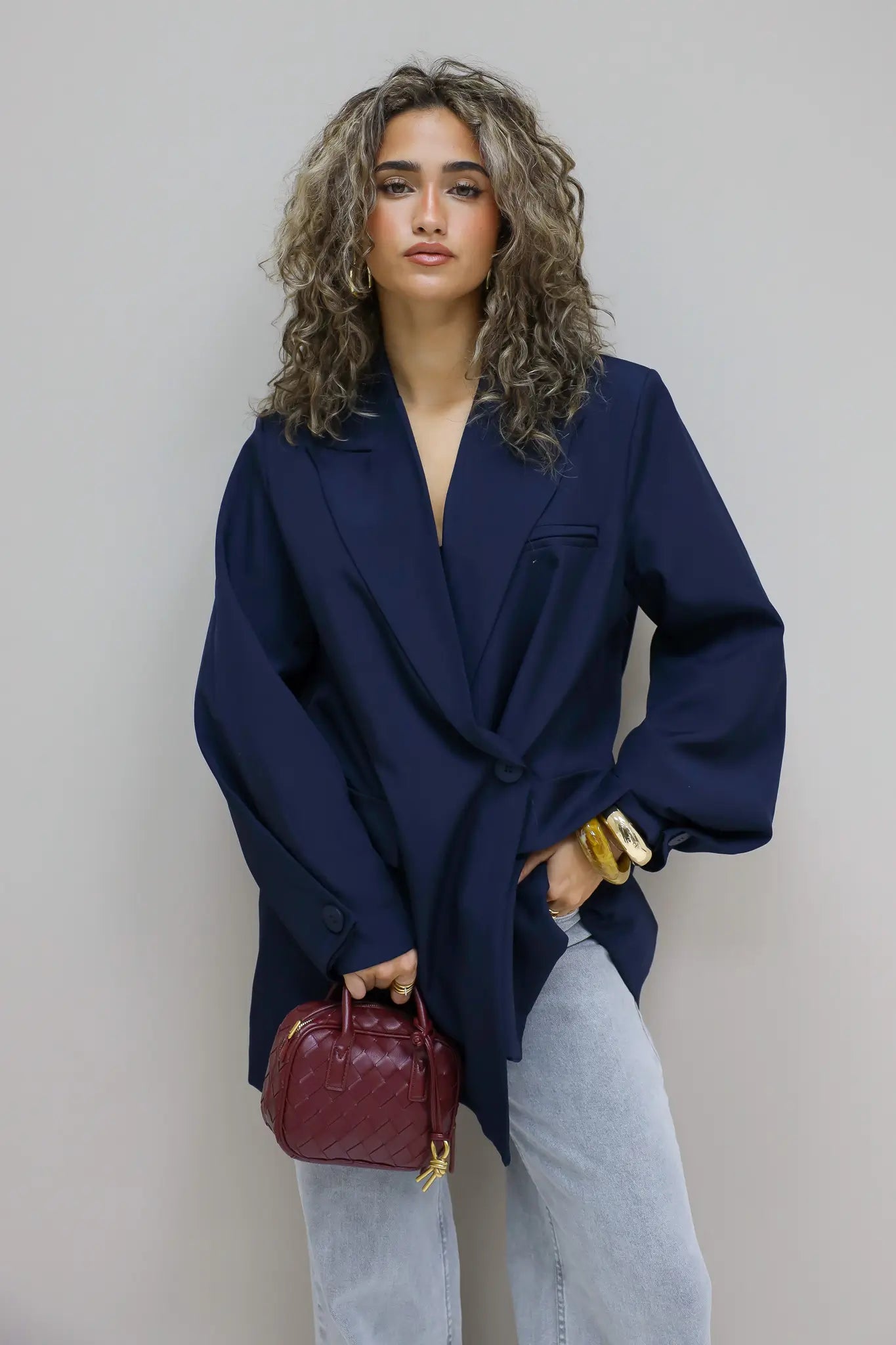 vrouw met krullen draagt donkerblauwe oversized blazer en kijkt recht vooruit tegen grijze muur
