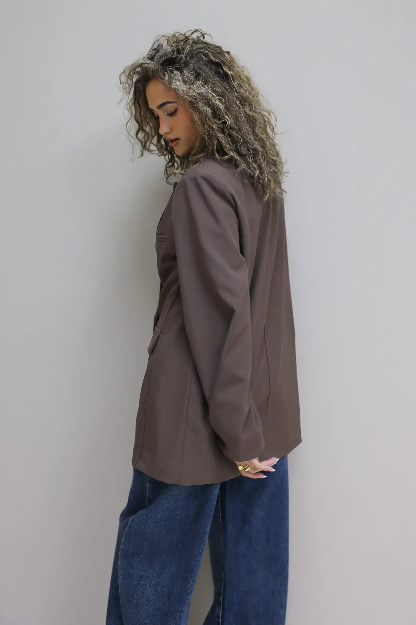 vrouw met krullen draagt donkerbruine oversized blazer en jeans tegen grijze muur