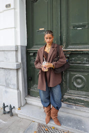 vrouw met donkerbruine oversized blazer, laarzen en jeans poseert bij stenen muur