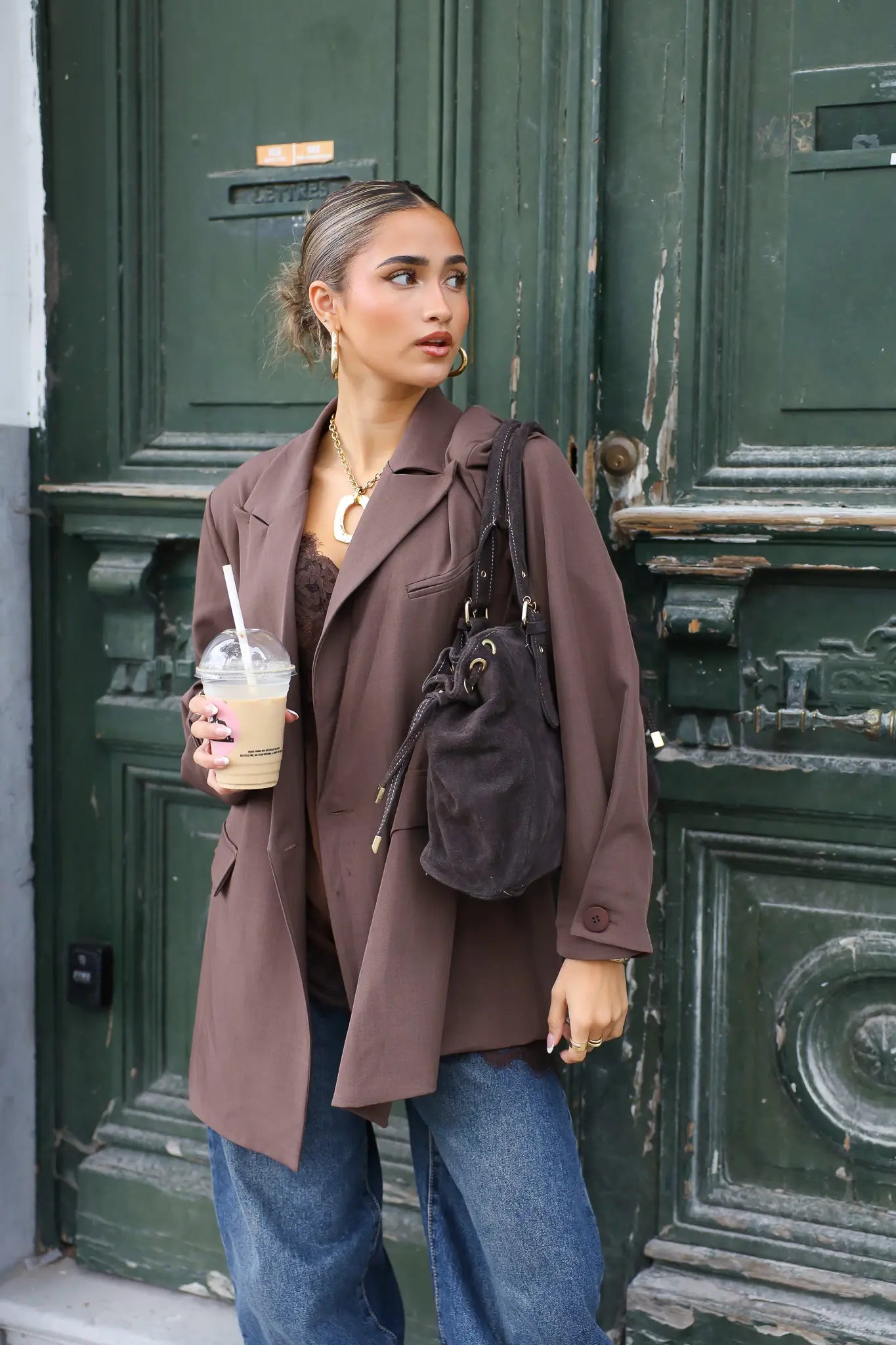 vrouw draagt donkerbruine oversized blazer met jeans en tas aan arm voor groene deur
