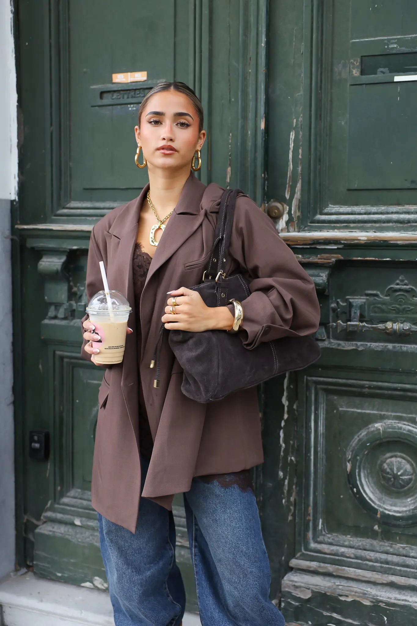 vrouw met donkerbruine oversized blazer en jeans kijkt recht vooruit bij groene deur