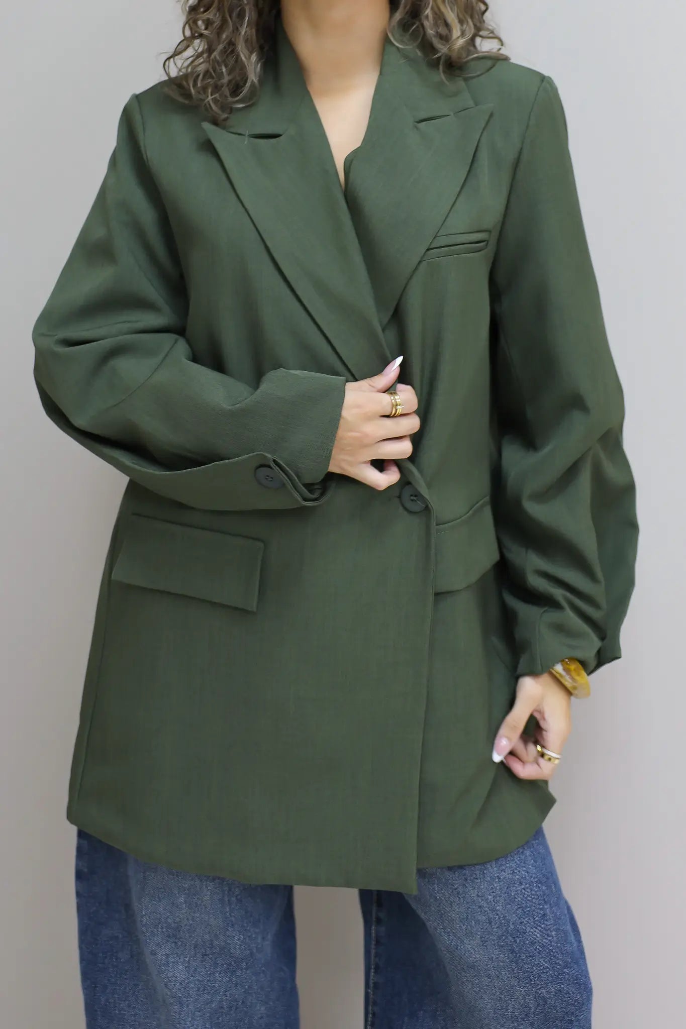 vrouw met krullen draagt groene oversized blazer en houdt revers vast tegen grijze muur