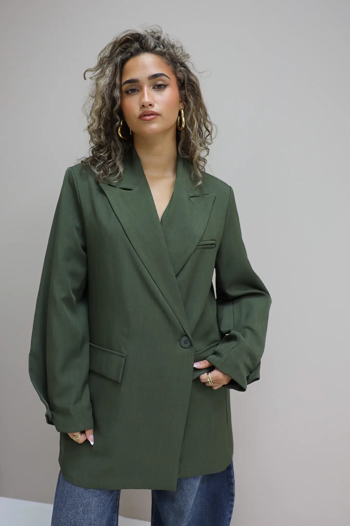 vrouw kijkt recht vooruit met groene oversized blazer en jeans tegen grijze achtergrond