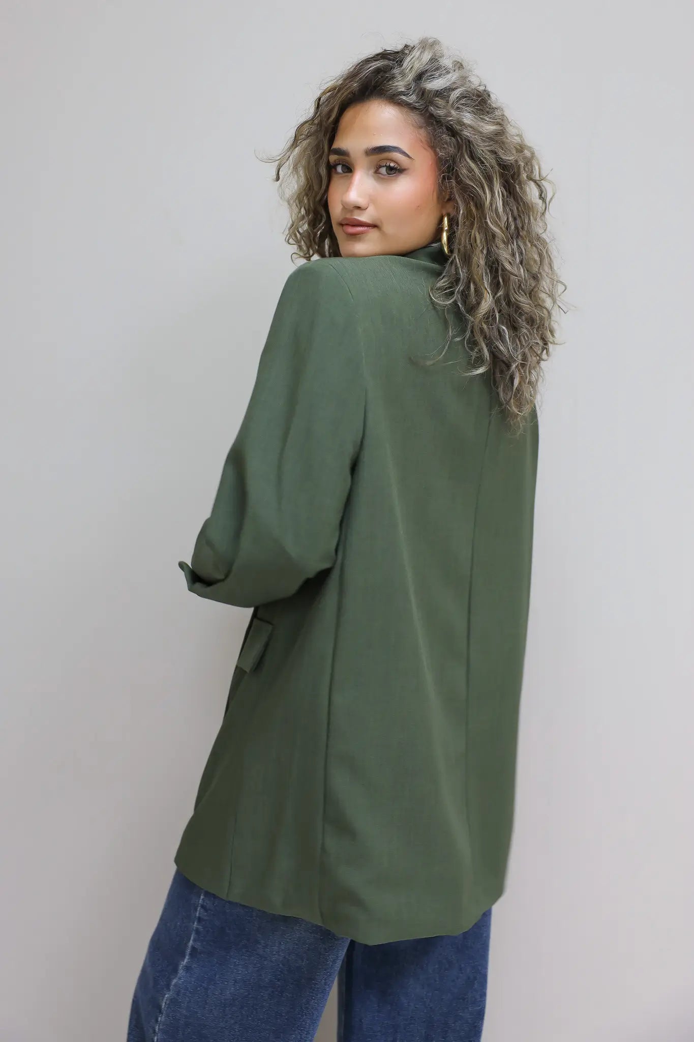 zijaanzicht van vrouw met groene oversized blazer en jeans tegen grijze muur