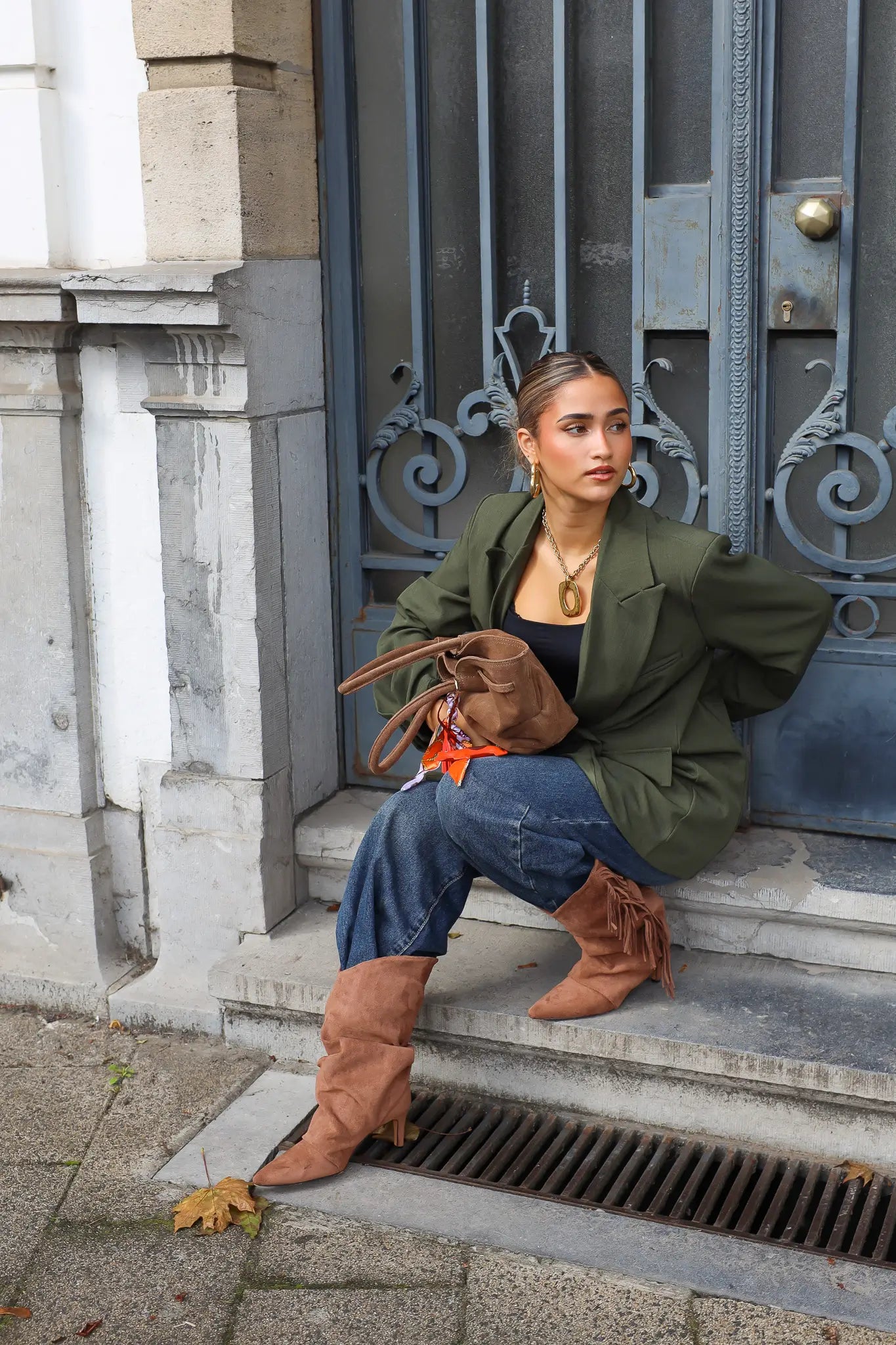 vrouw met groene oversized blazer en bruine laarzen zit op stoep bij blauwe deur