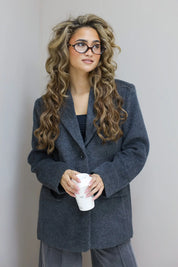 Model met donkergrijze wolblazer, kijkt recht vooruit, handen in zakken