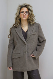 Model poseert tegen muur, taupe blazer los gedragen