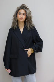 vrouw met zwarte oversized blazer en lichte jeans tegen grijze achtergrond binnen