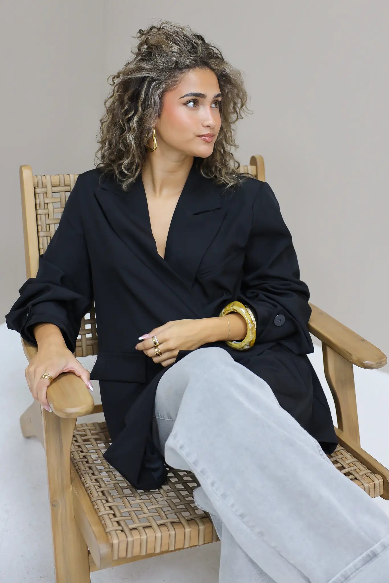 vrouw met krullen draagt zwarte oversized blazer open zittend op houten stoel binnen