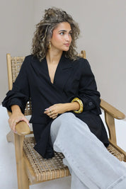 vrouw met krullen draagt zwarte oversized blazer open zittend op houten stoel binnen