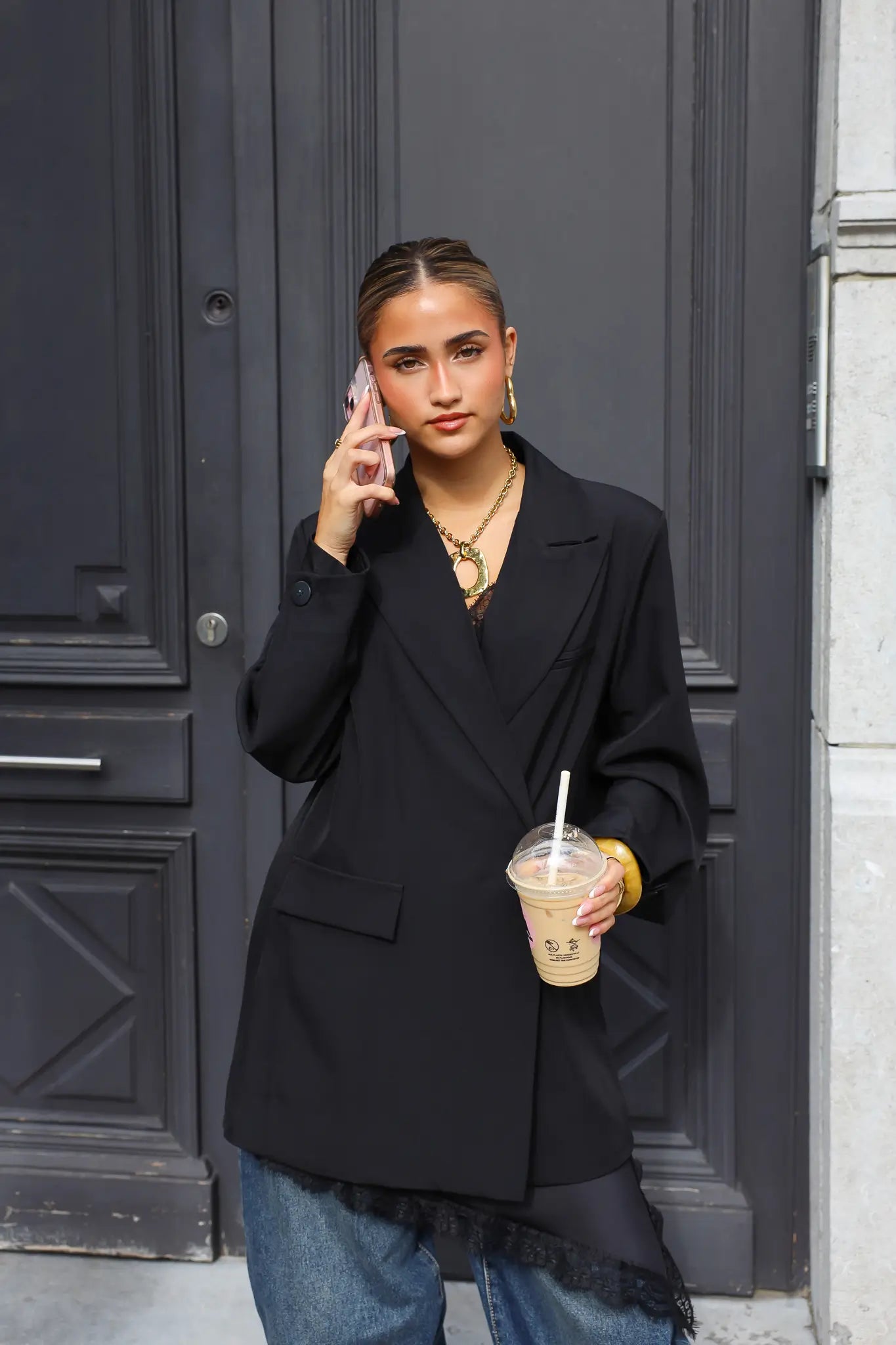 vrouw draagt zwarte oversized blazer met gouden ketting en iced coffee in de hand voor donkere deur