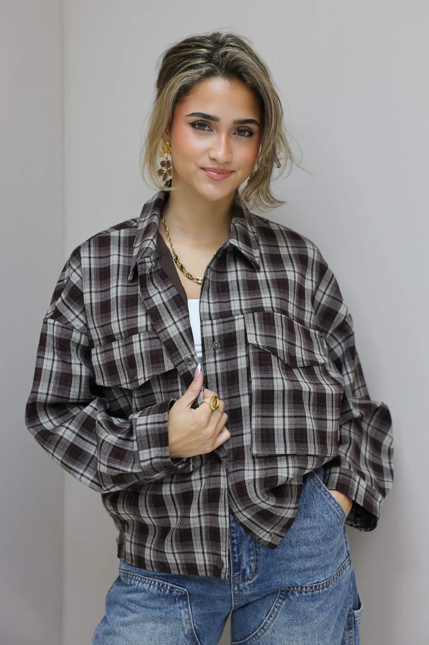 vrouw met bruine geruite blouse en jeans poseert tegen grijze muur