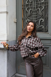 vrouw draagt bruine geruite blouse met bruine broek en poseert buiten tegen muur