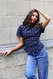 Model draagt een donkerblauwe geruite blouse met korte mouwen, knoopdetails en getailleerde pasvorm, gecombineerd met een lichtblauwe jeans.