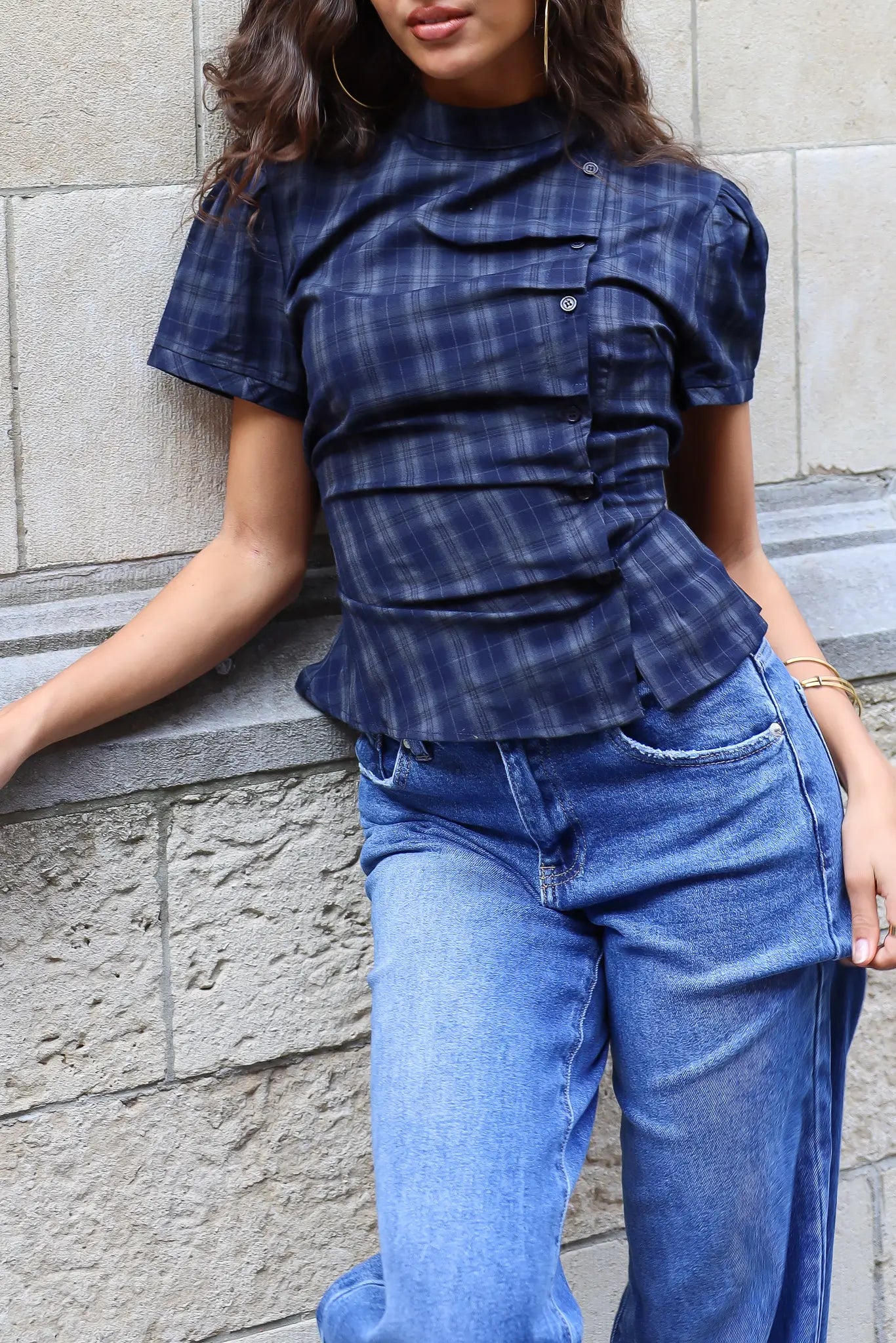 Detail van een donkerblauwe geruite damesblouse met korte mouwen, asymmetrische knoopsluiting en uitlopende taille, gedragen op een blauwe jeans.