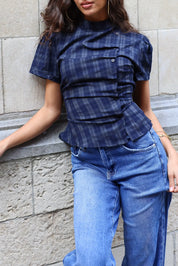 Detail van een donkerblauwe geruite damesblouse met korte mouwen, asymmetrische knoopsluiting en uitlopende taille, gedragen op een blauwe jeans.