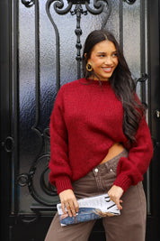 Model draagt bordeaux rode trui met bruine broek en witte clutch bij zwarte deur