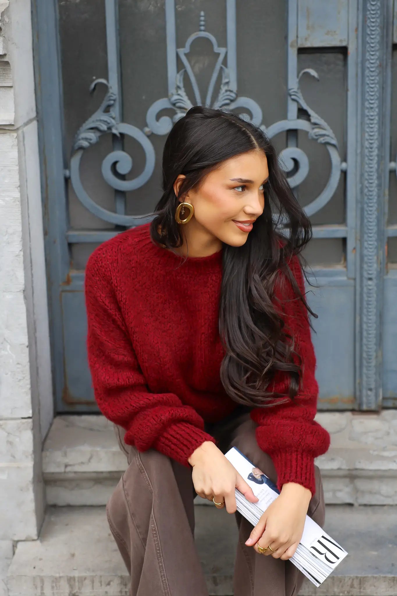 Model zit op traptreden in bordeaux rode trui met bruine broek en koffiebeker