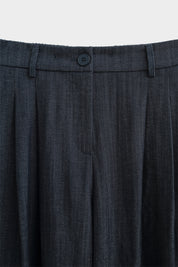 Close-up van de taille van een donkerblauwe wide fit broek met riemlussen, knoopsluiting en plooien aan de voorkant.