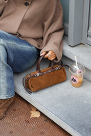 Bruine suède handtas met donkerbruine leren details en zilverkleurige gespen naast een blauwe jeans en bruine jas