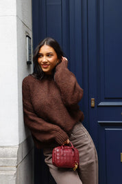 Model draagt bruine fluffy trui met bruine broek en rode tas bij blauwe deur