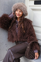 Model zit met bruine fur jas, taupe muts en tas met studs