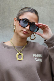 model met bruine zonnebril en goudkleurige oorbellen poseert buiten tegen grijze muur