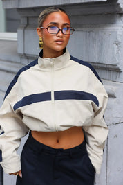 Camel track jacket met donkerblauwe streep over de borst en hoge kraag gedragen met een zonnebril