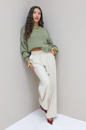 Model leunt tegen muur in cremekleurige pantalon met groene trui