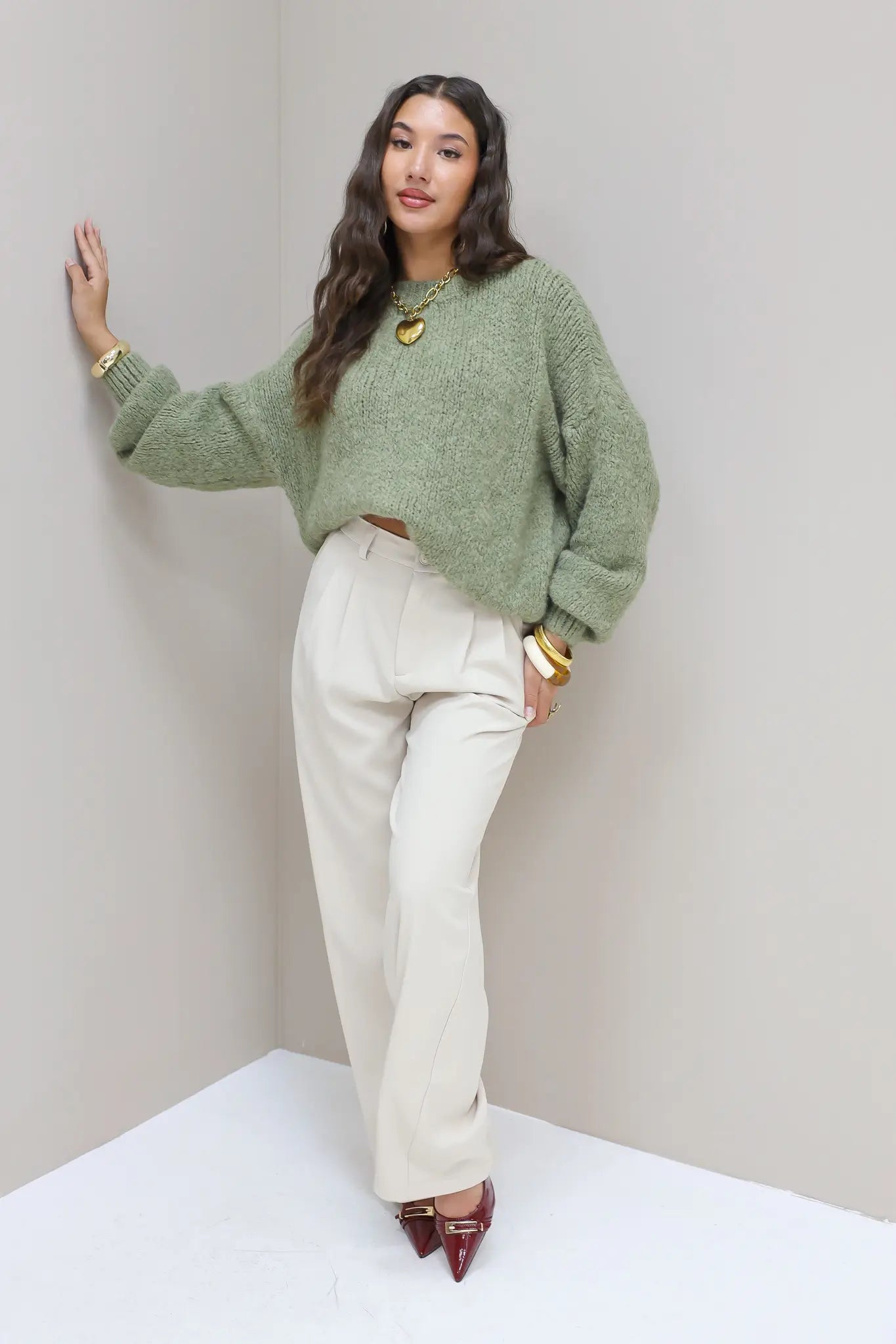 Model poseert in hoek met groene trui en cremekleurige pantalon