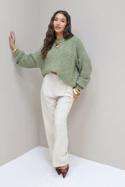 Model poseert in hoek met groene trui en cremekleurige pantalon