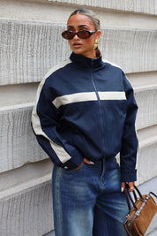 Donkerblauw track jacket met beige streep over de borst en hoge opstaande kraag gedragen bij een blauwe jeans