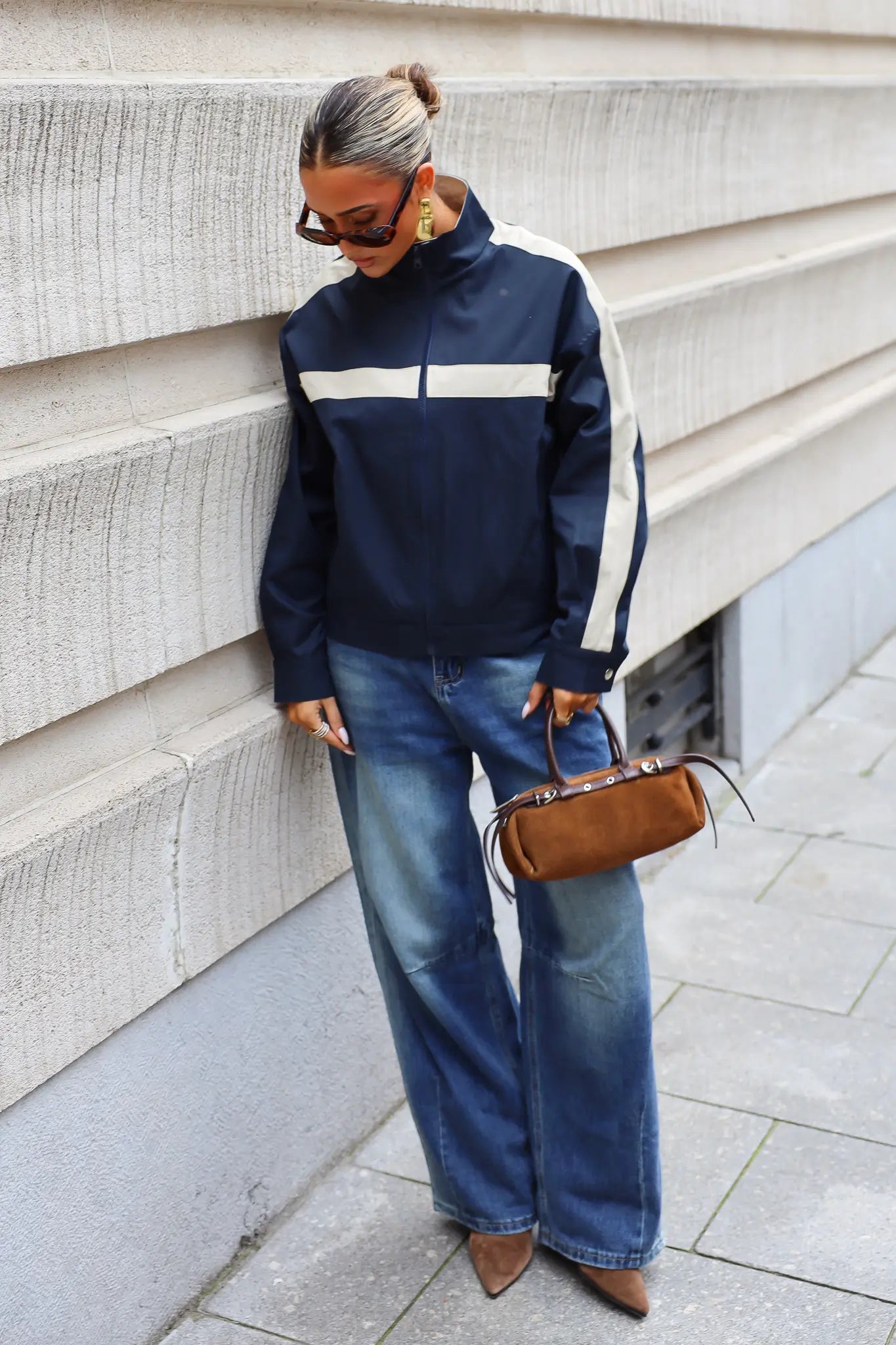Donkerblauwe track jas met ritssluiting en contrasterende beige strepen gedragen bij een blauwe wide leg jeans en bruine handtas