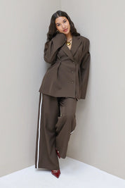 Model leunt in hoek tegen muur in donkerbruine blazer met pantalon met witte bies